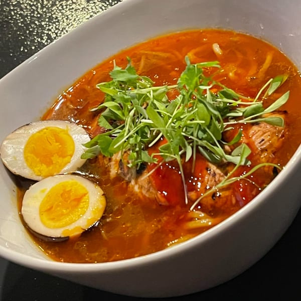 A bowl of rich red ramen with noodles, meat, two halved boiled eggs, and fresh greens.