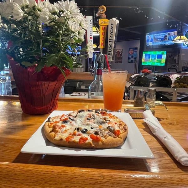A small pizza, orange drink, and white flowers on a wooden bar with beer taps and TVs.