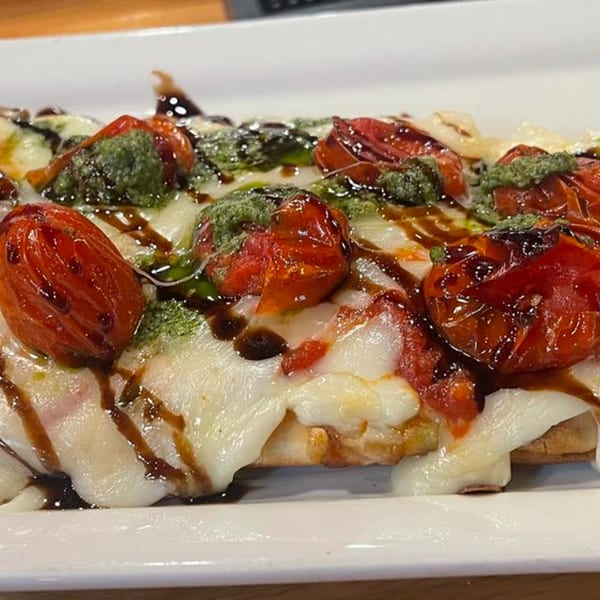 Flatbread pizza with melted mozzarella, roasted tomatoes, pesto, and balsamic glaze on a white plate.